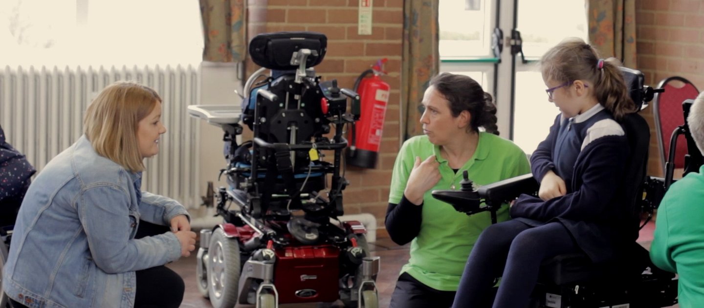 A Whizz Kidz clinician speaks with a young wheelchair user and her mother