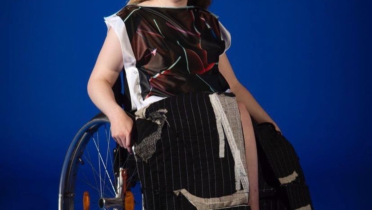 Caitlyn Fulton in a wheelchair, wearing a dark red top with white vertical edges, and thin light blue rectangles at irregular angles. Her skirt is black with white pinstripes and she is wearing black shoes. She has a neutral expression and her right hand