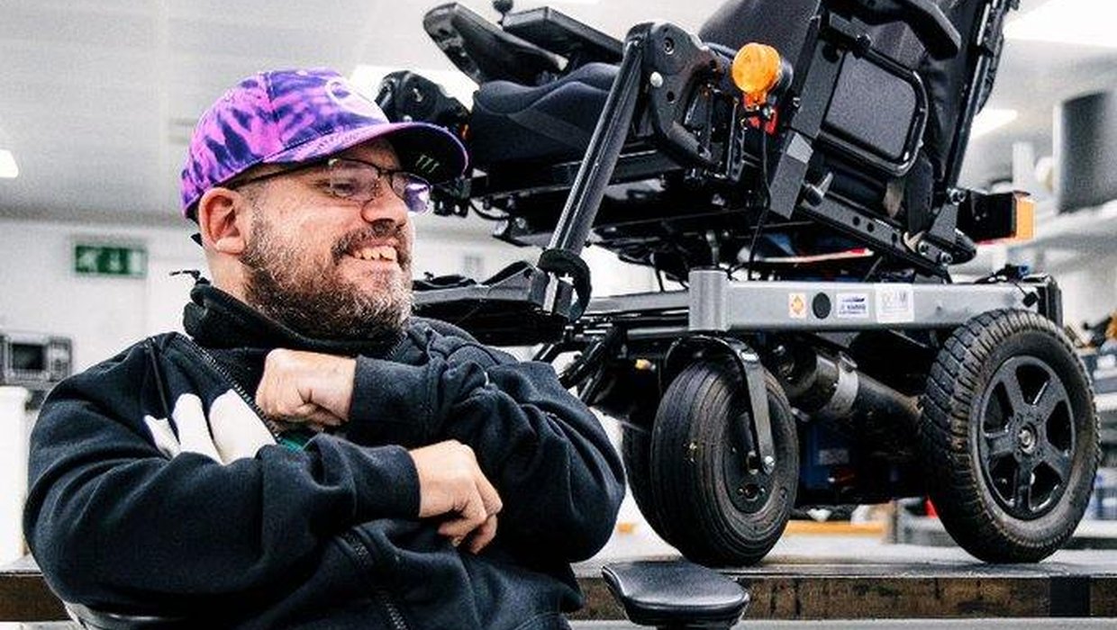 Adam Stanton-Wharmby wearing a purple baseball cap, black zipped top and jeans. Behind him on a  raised platform is his F1 enhanced wheelchair.