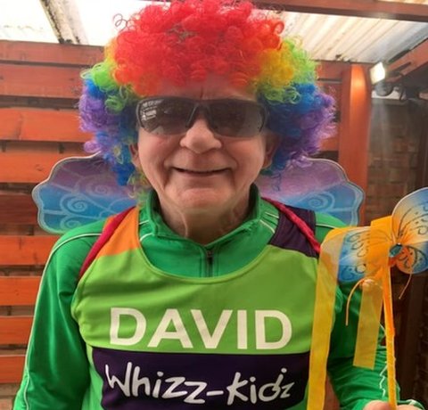 David is wearing a green Whizz Kidz running vest, with a colourful wig, wings and cool magic wand