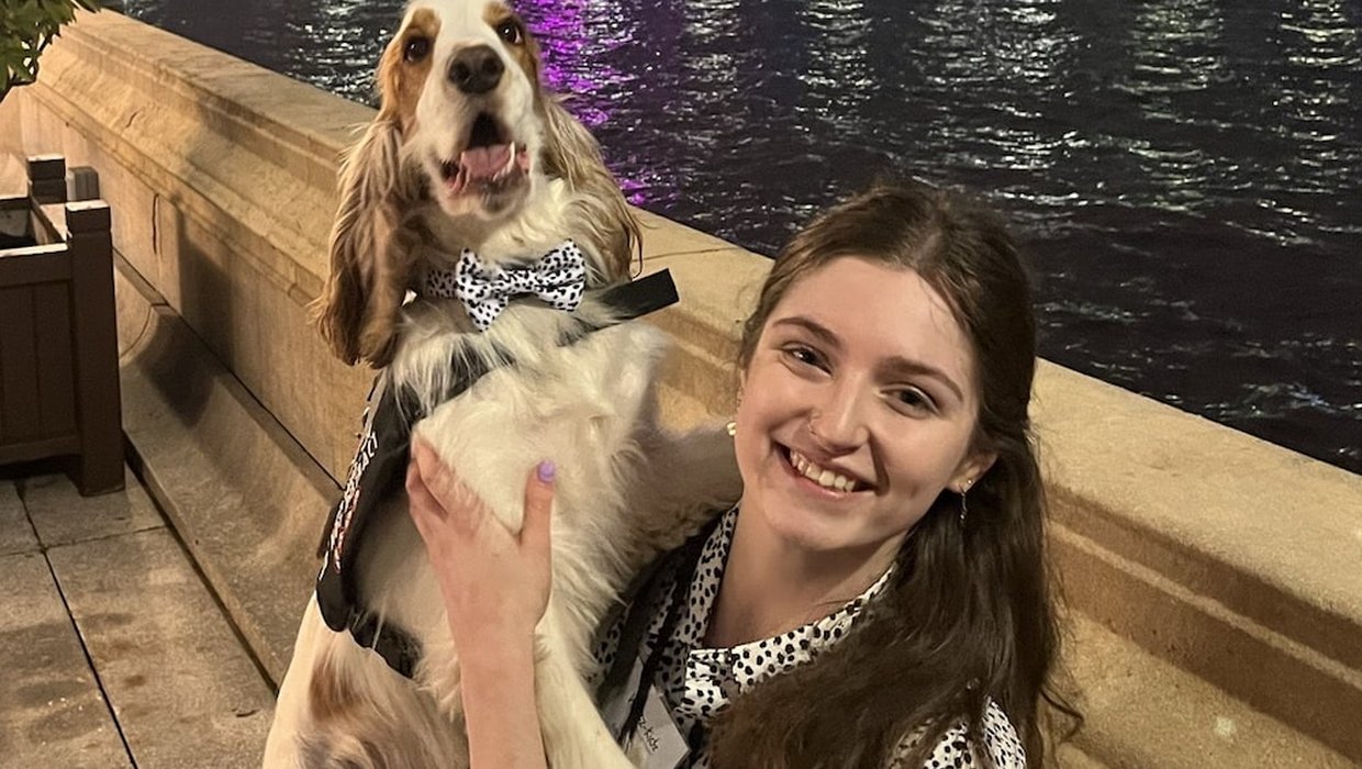 Ella with Moose at the Houses of Parliament. Moose is wearing a spotty bow tie that matches Ella's dress. They are on the terrace by the river Thames at night.