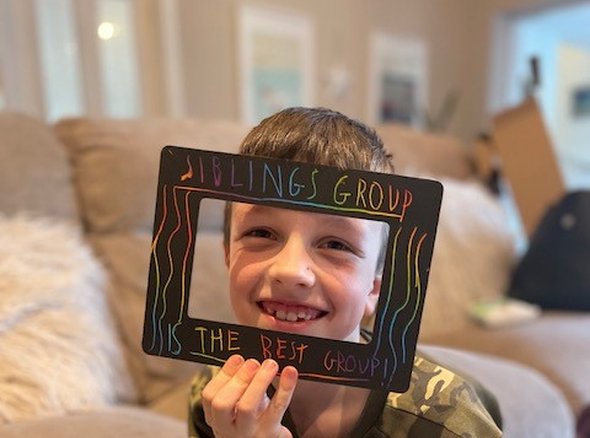A child holds a frame in front of him that reads 'Sibling Group the best group'